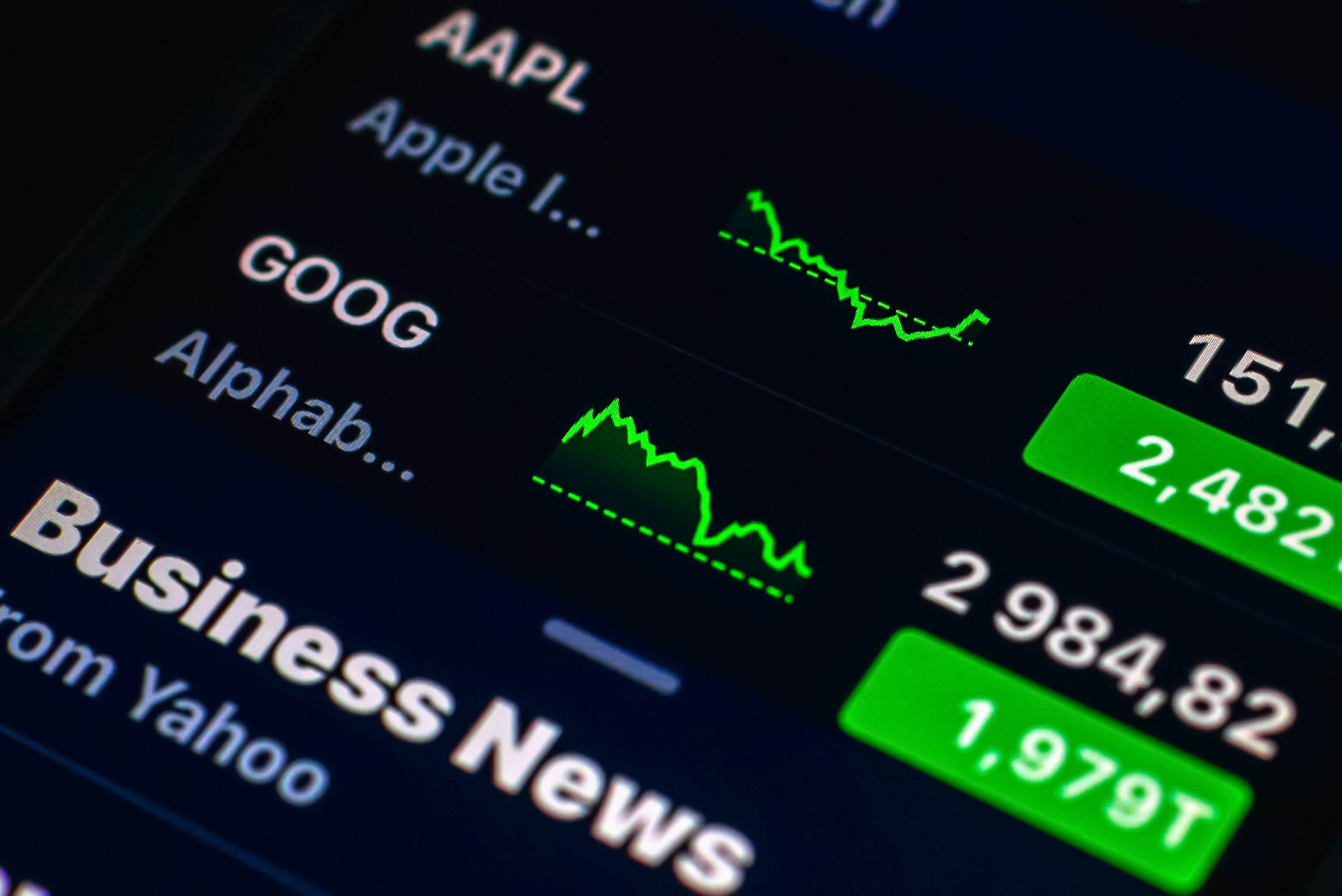 A close-up of a digital stock market display showing tickers for Apple (AAPL) and Alphabet (GOOG) alongside green performance line graphs.